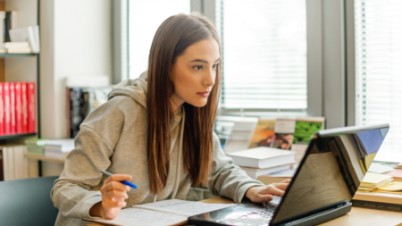 Eine junge Frau beugt sich ihrem Laptop entgegen. Sie hält einen Stift in der Hand und macht sich Notizen.